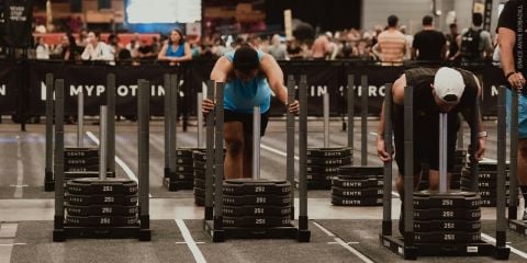 Bei einem gut besuchten Indoor-Fitnesswettbewerb schieben zwei Sportler mit Gewichten beladene Schlitten. Im Hintergrund beobachten Zuschauer das Geschehen hinter Absperrungen.