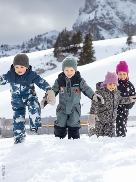 Vier Kinder in Winterkleidung spielen und lächeln im Schnee, im Hintergrund sind schneebedeckte Berge und Bäume zu sehen.
