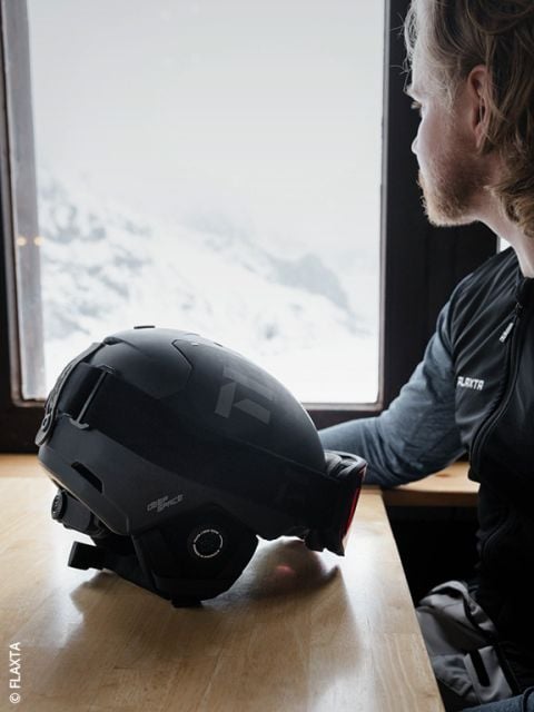 Eine Person mit hellem Haar und Bart sitzt an einem Holztisch am Fenster und blickt nach draußen auf verschneite Berge. Ein schwarzer Skihelm liegt vor ihnen auf dem Tisch.