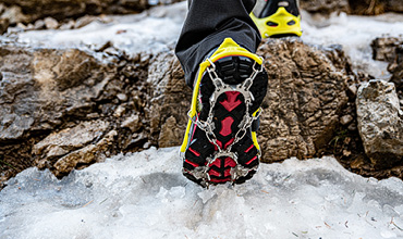 Nahaufnahme einer Person, die auf eisbedeckten Felsen wandert. Sie trägt leuchtend gelbe und schwarze Schuhe, die mit Metallsteigeisen für den Halt ausgestattet sind. Die Szene betont Winteraktivitäten im Freien.